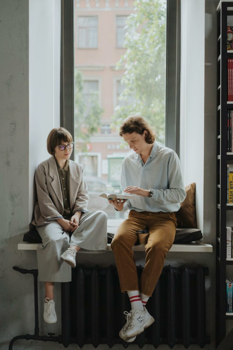 Two young adults enjoy a book in a cozy indoor setting by the window.